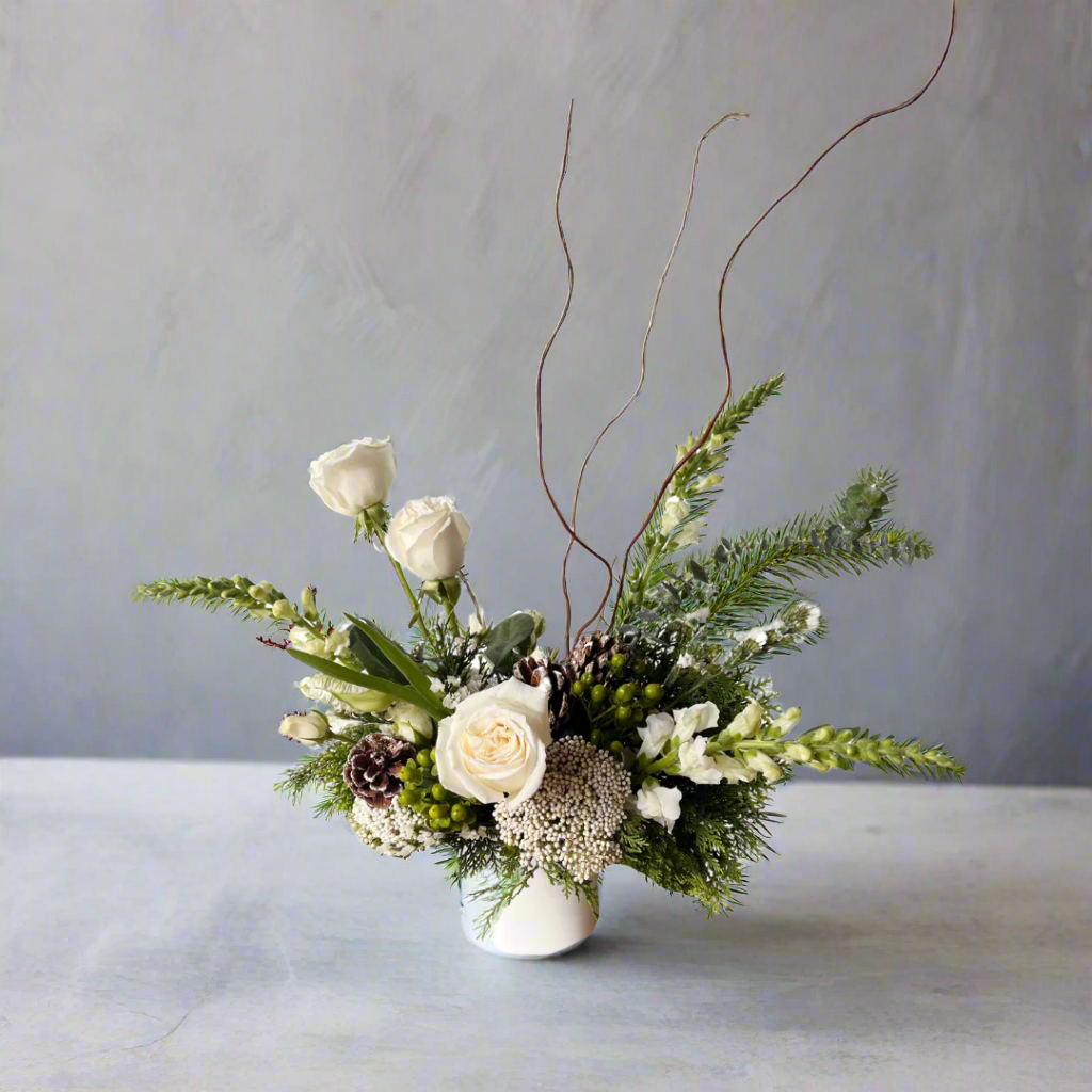 Floral arrangement with white roses, greenery, and pinecones on a light surface.